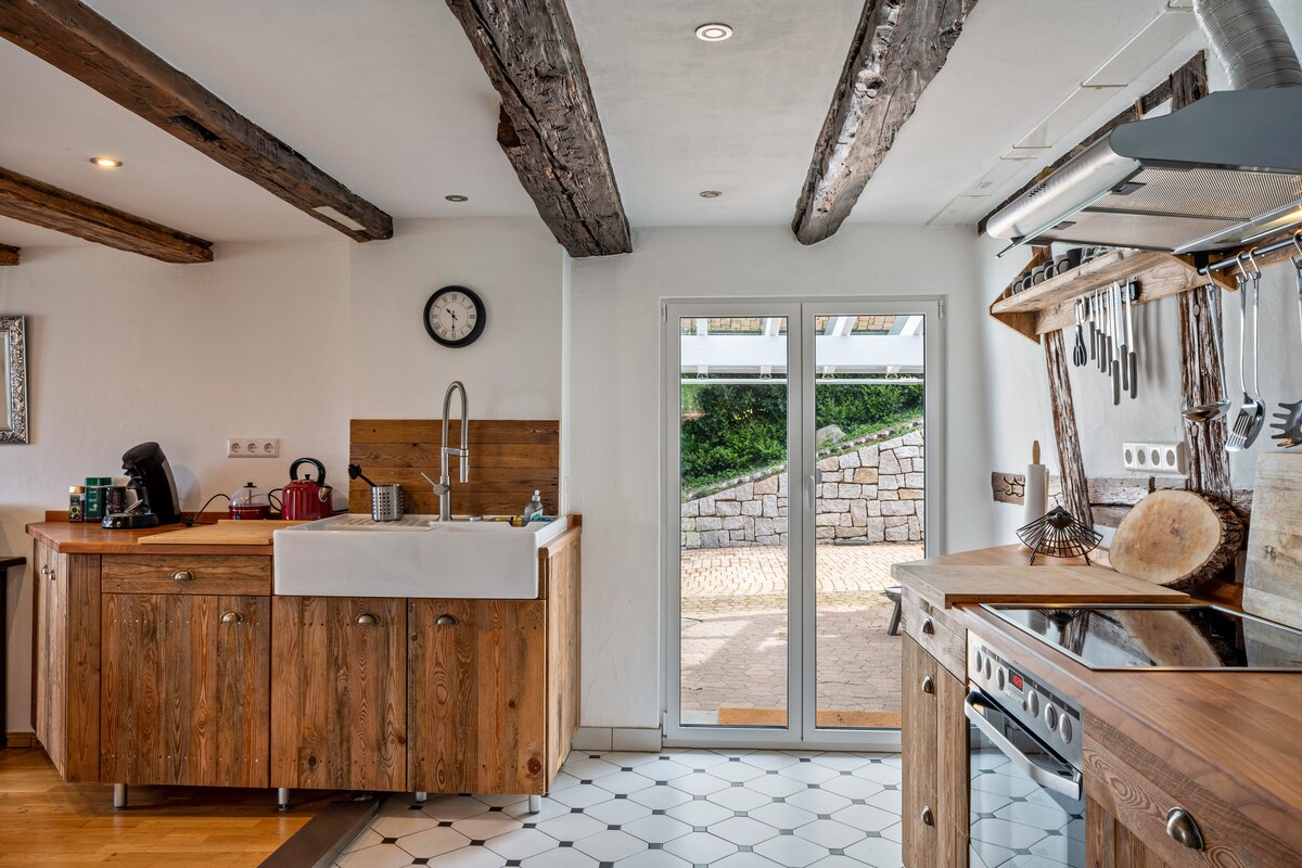 The kitchen features wooden cabinetry and a farmhouse sink, complemented by exposed wooden beams above. A large glass door opens to the outdoor area, while a clock and modern appliances are visible. A stylish countertop provides additional workspace.