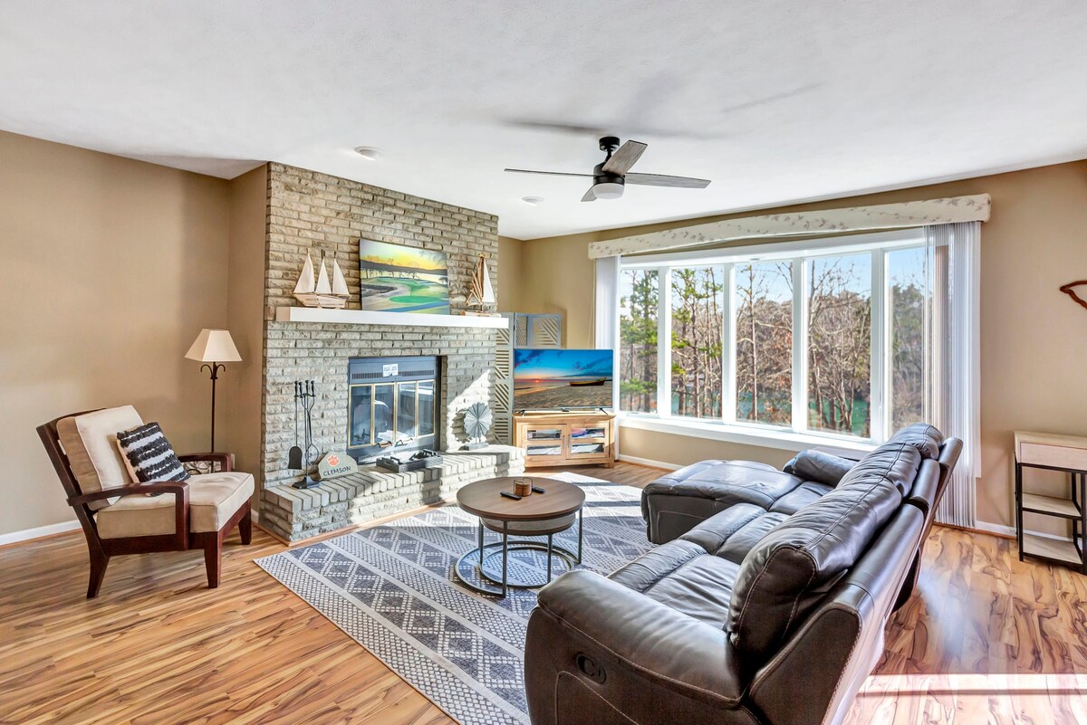 A spacious living area is highlighted by a stone fireplace, complemented by a large window offering views of the surrounding greenery. Comfortable seating is arranged around a central coffee table, with a subtle area rug adding texture to the hardwood floor.
