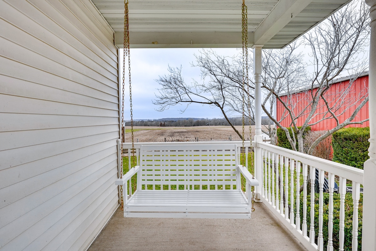 A welcoming porch swing is suspended from a white canopy, providing an unobstructed view of the surrounding fields. The gentle landscape features a blend of greenery and farmland, with a nearby red barn visible in the distance under a cloudy sky.