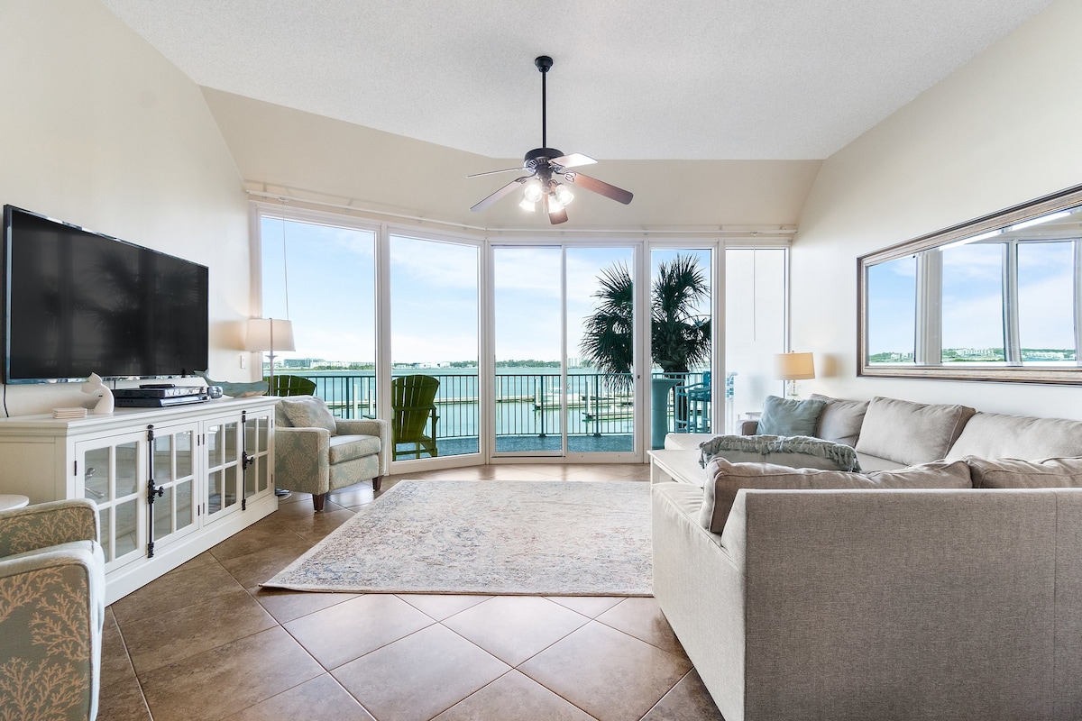 A welcoming living area features a light-colored sectional sofa and an armchair, complemented by a soft area rug. A large flat-screen TV is mounted on the wall, and expansive windows provide stunning views of the water and surrounding landscape.