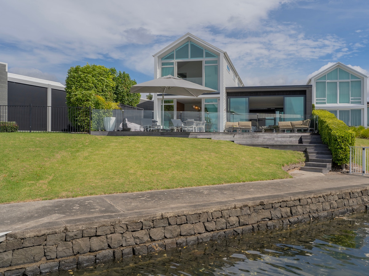 The exterior of Villa Viti features a well-maintained lawn leading to the waterfront. The modern structure boasts large glass sliding doors, offering a seamless connection to the outdoor entertaining area, shaded by a large umbrella. Stone walls border the pristine waterway.