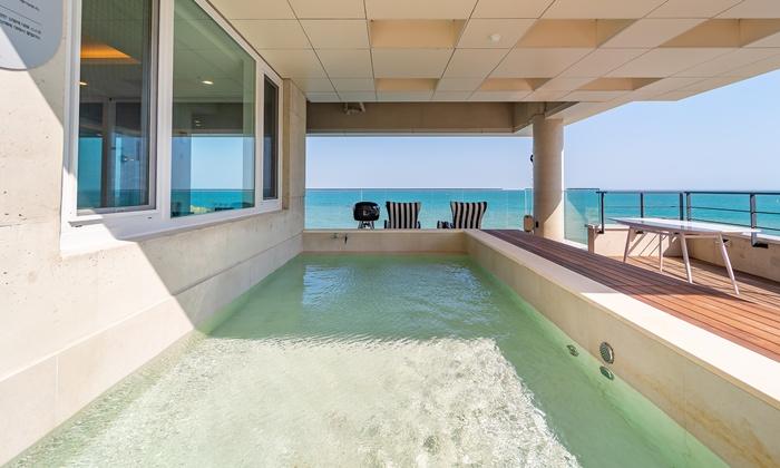 A private pool is set against a backdrop of blue waters, with a deck featuring striped lounge chairs nearby. Large windows provide ample sunlight, creating a serene atmosphere. A barbecue grill is positioned conveniently for outdoor cooking.