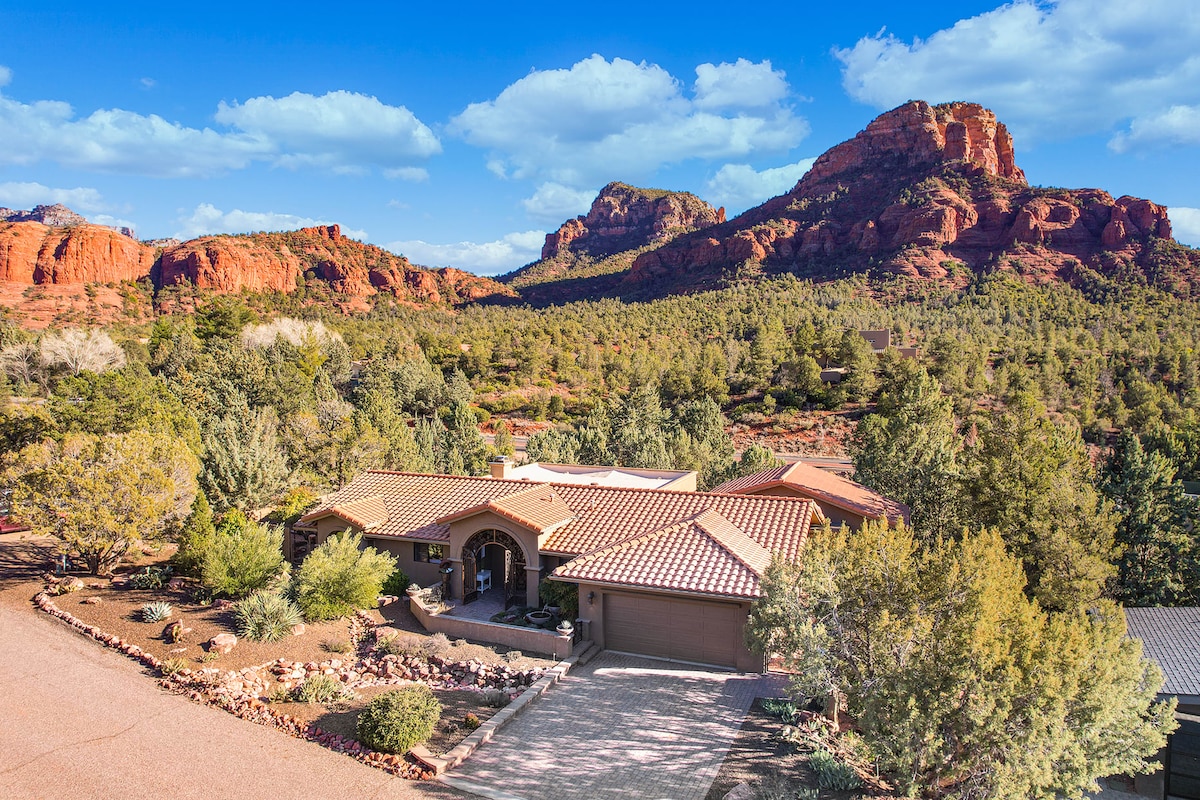 A spacious villa is set against a stunning backdrop of red rock formations and greenery. The structure features a terracotta tile roof and a landscaped entrance. The surrounding landscape showcases a blend of trees and rocky outcrops, inviting a connection to nature.