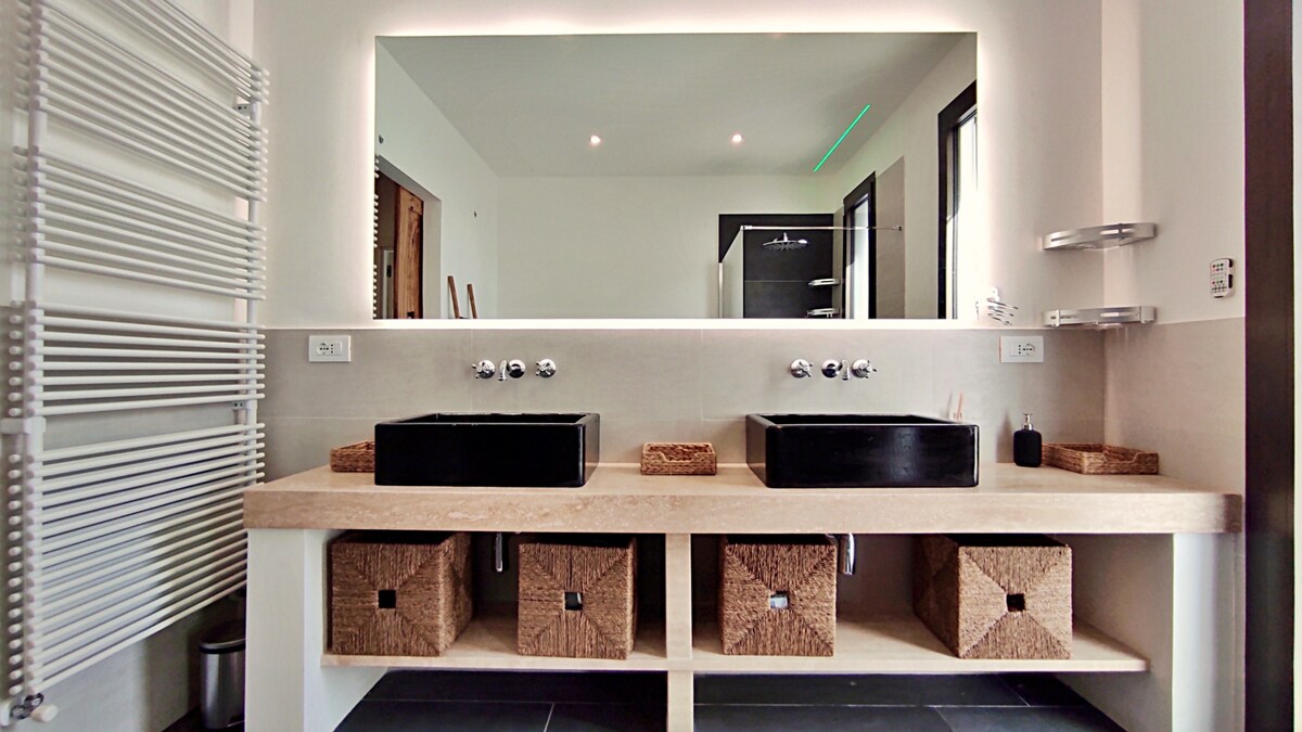 A spacious bathroom is equipped with a long countertop featuring dual black vessel sinks. Below the countertop, woven storage baskets provide organized space for essentials. A full-length mirror reflects soft lighting, and dark tiles enhance the modern design. The shower area is visible in the background.