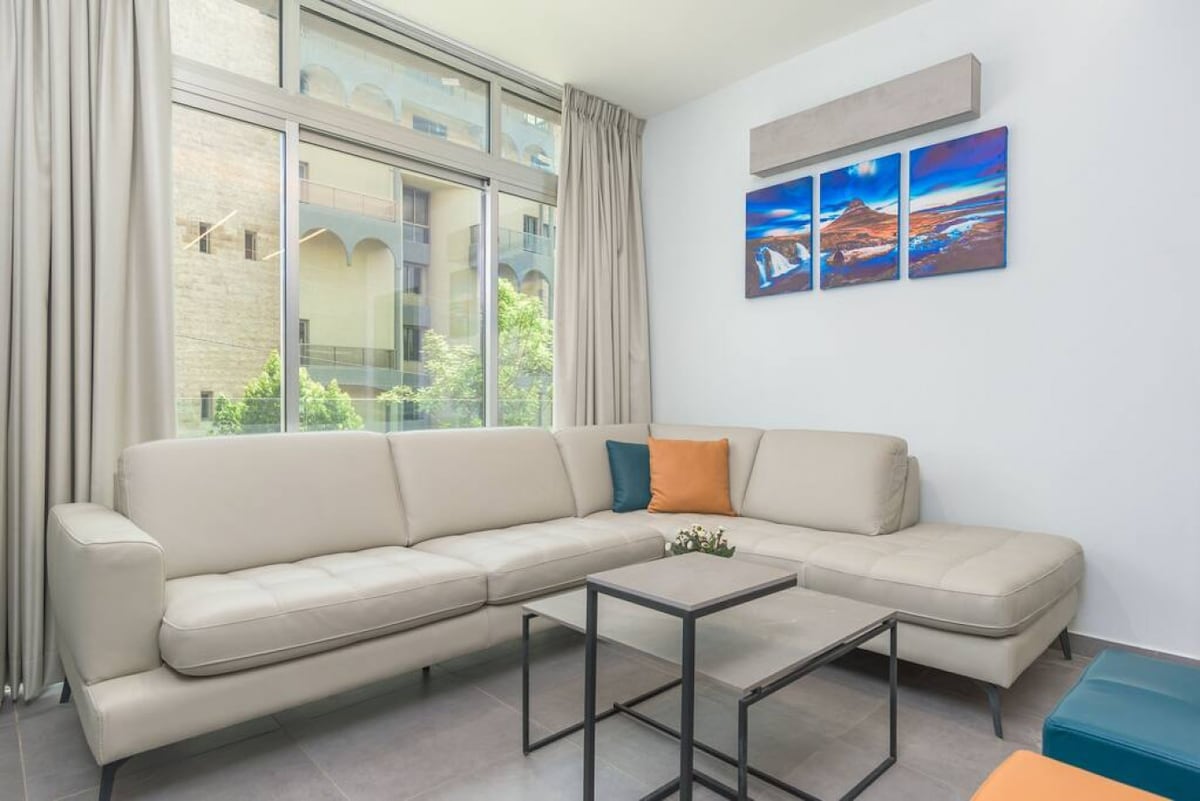 A light-filled living area features a modern sectional sofa in muted tones, complemented by decorative cushions. A sleek coffee table sits in front, while framed artwork adorns the wall, adding a splash of color. Expansive windows offer views of the surrounding greenery.