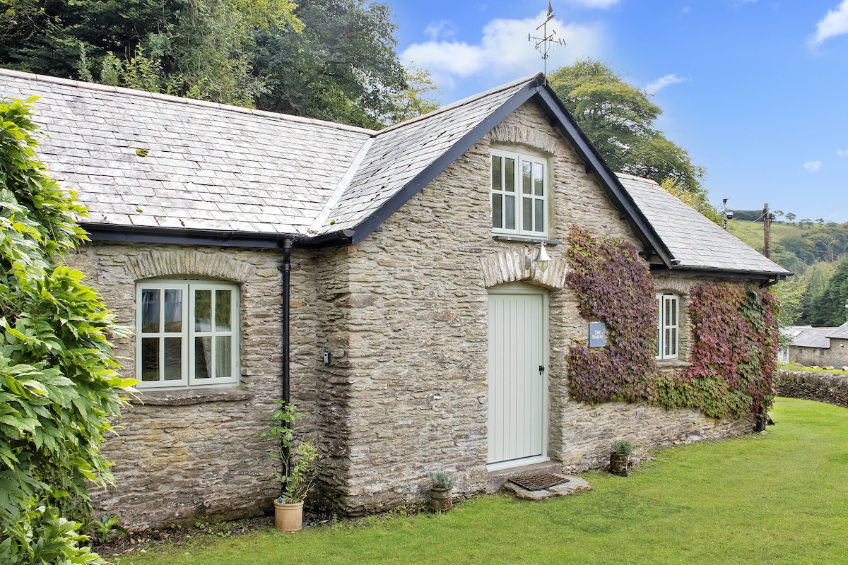 A traditional stone cottage is shown with a slate roof and a single, central entrance. Vines climb the exterior wall, with plants placed near the entrance. Natural greenery surrounds the property, enhancing the cottage's charming rural aesthetic.