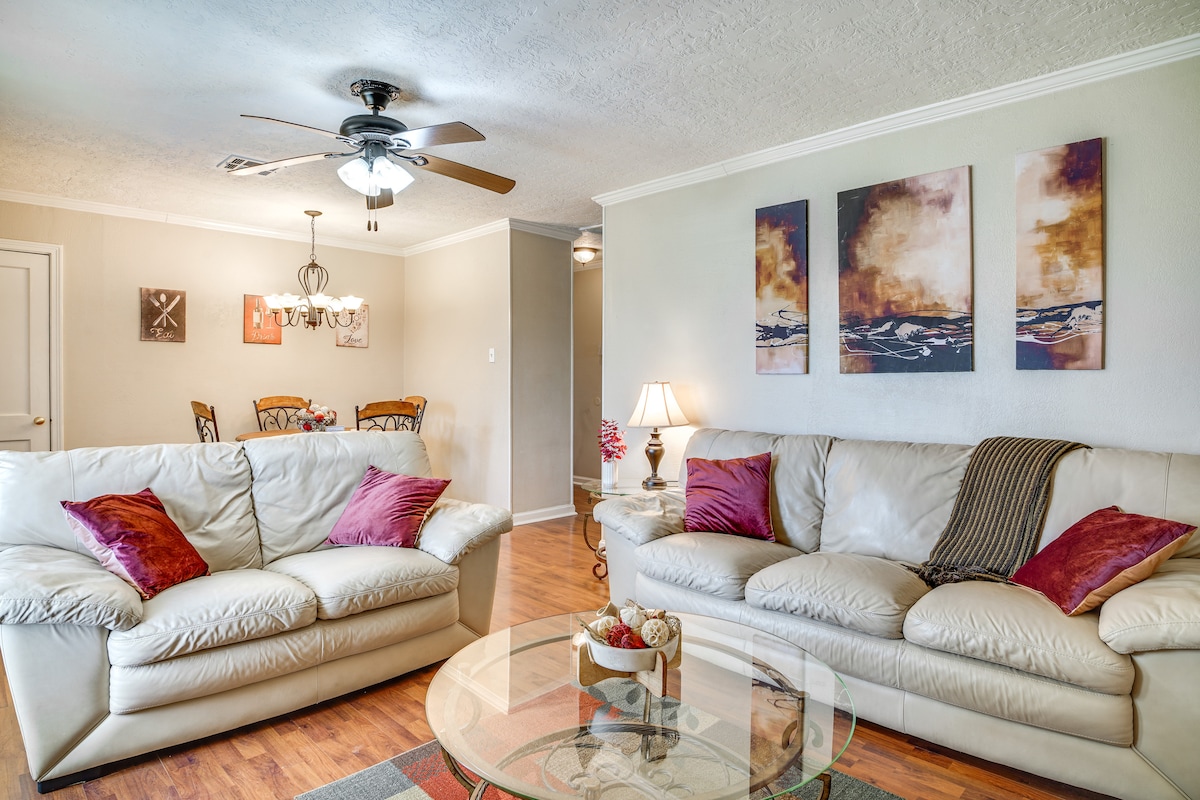 A living area is furnished with two light-colored sofas embellished with throw pillows. A decorative glass coffee table sits in the center, while a ceiling fan provides comfort. Artwork adorns the wall, and a dining area is visible in the background.