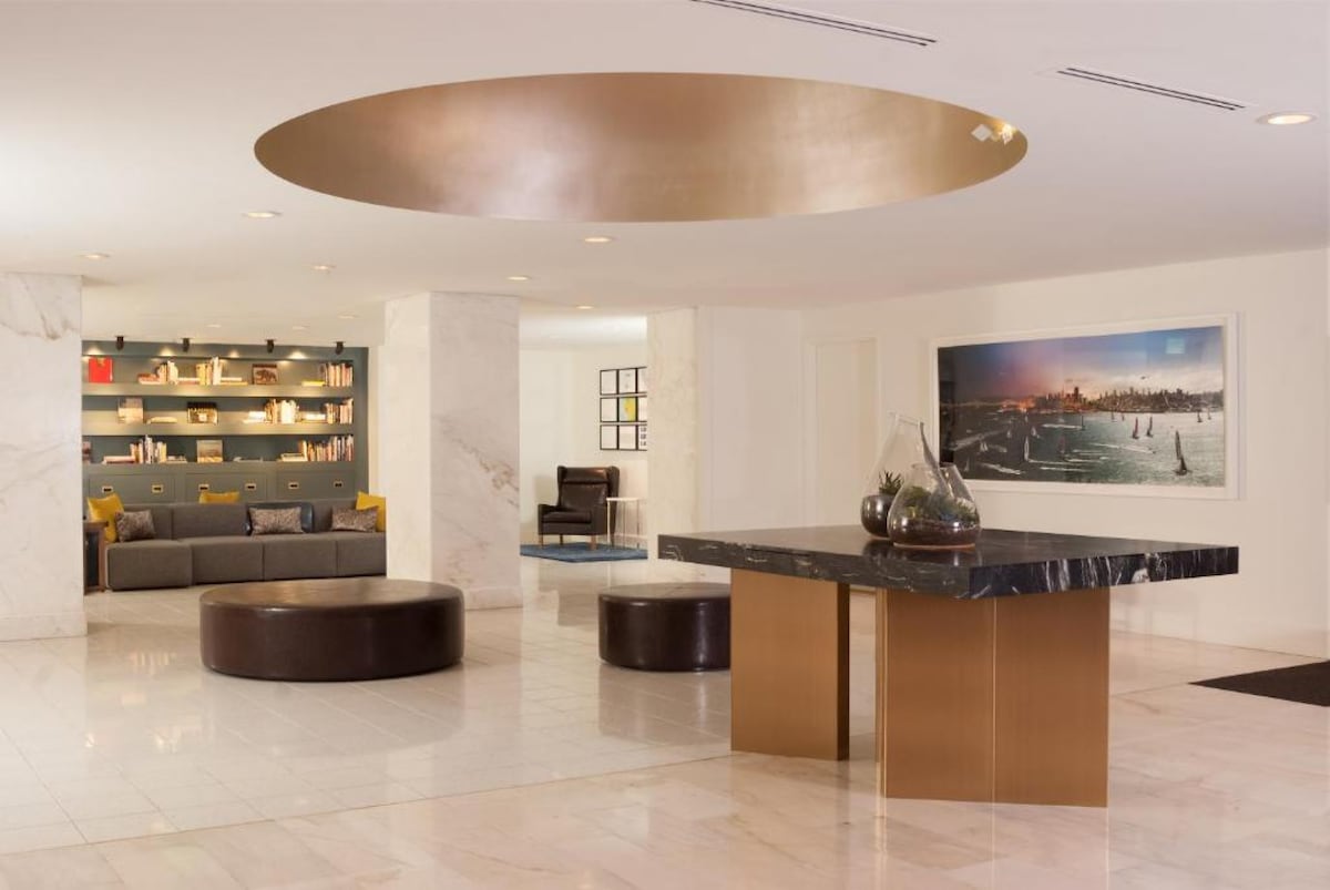 The lobby features a spacious layout with polished marble flooring and a large circular ceiling detail. A modern reception desk crafted from dark stone stands at the center. Comfortable seating areas with gray sofas and decorative shelves filled with books create a welcoming atmosphere.