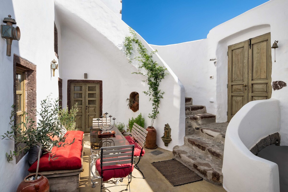 A charming patio area is surrounded by white walls, featuring a wooden table accompanied by red cushions on benches. A few plants add greenery, while traditional pottery decorates the space. Stairs lead to an upper level, creating a cozy, inviting atmosphere.