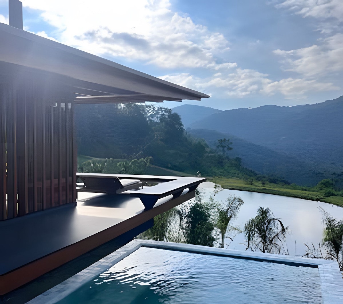 A modern structure extends over the serene lake, featuring a spacious deck that provides sweeping views of the surrounding mountains. The calm water reflects the sky, while the greenery adds a natural touch to the tranquil setting.
