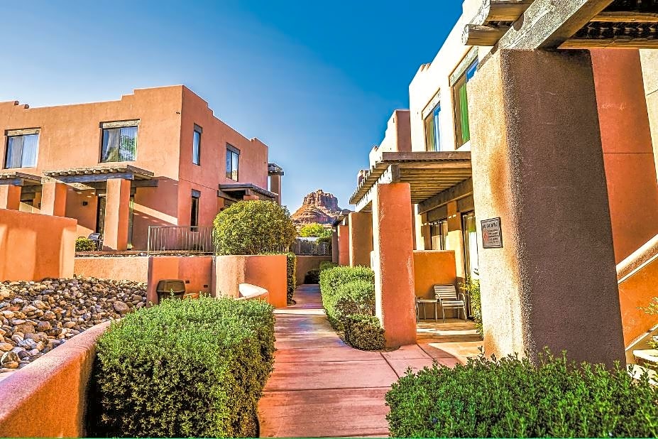 The image showcases a pathway lined with well-maintained green shrubs, leading past adobe-style buildings. Natural rock formations can be seen in the background under a clear blue sky, creating a harmonious blend with the surrounding landscape.