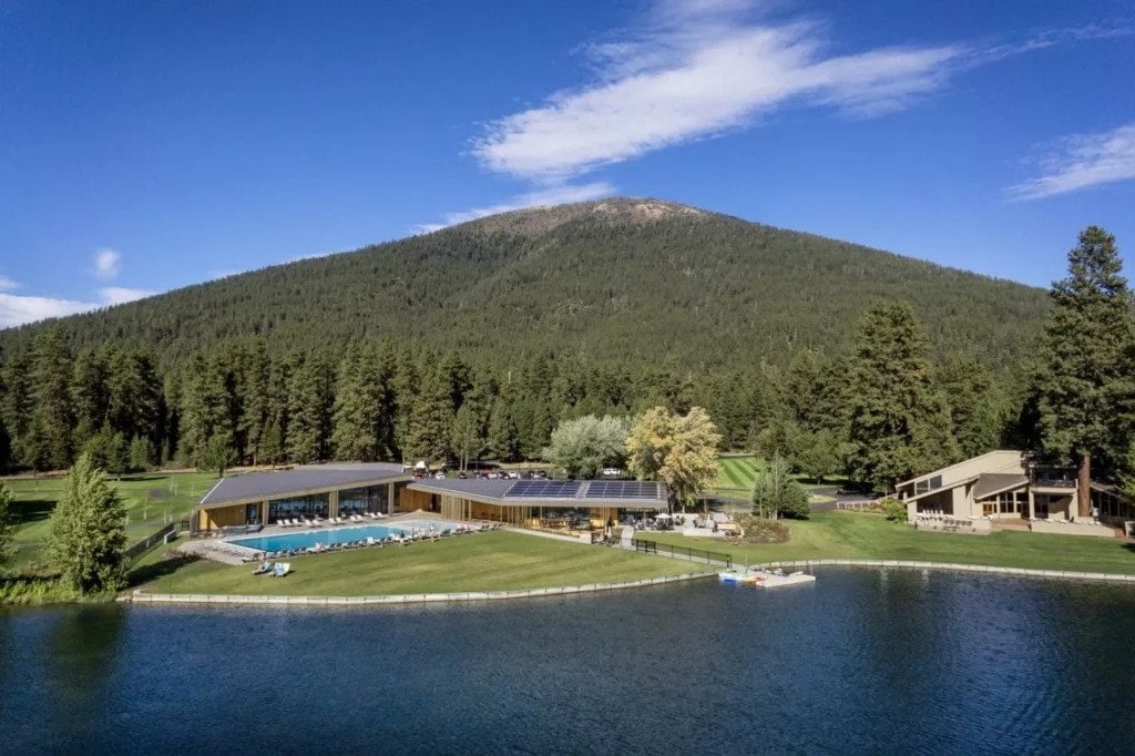 A scenic view captures the expansive landscape featuring a serene lake bordered by lush green lawns and dense forests. In the background, a mountain rises under a blue sky dotted with clouds. The modern structure with multiple rooftops includes an inviting pool area, complementing the natural beauty.
