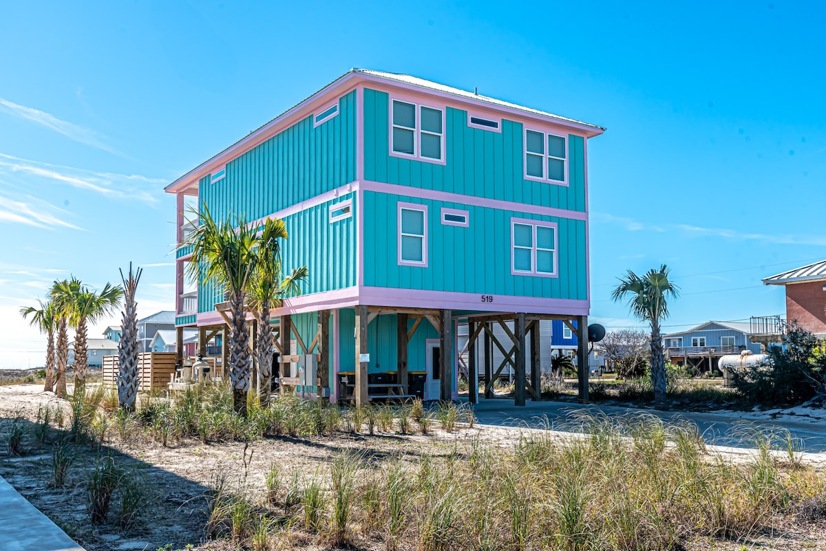 A two-story, custom-built home features a vibrant exterior in shades of blue and pink, complemented by palm trees and grassy landscaping. Elevated construction allows for parking underneath, and large windows provide ample light. The beach is accessible via a nearby path.