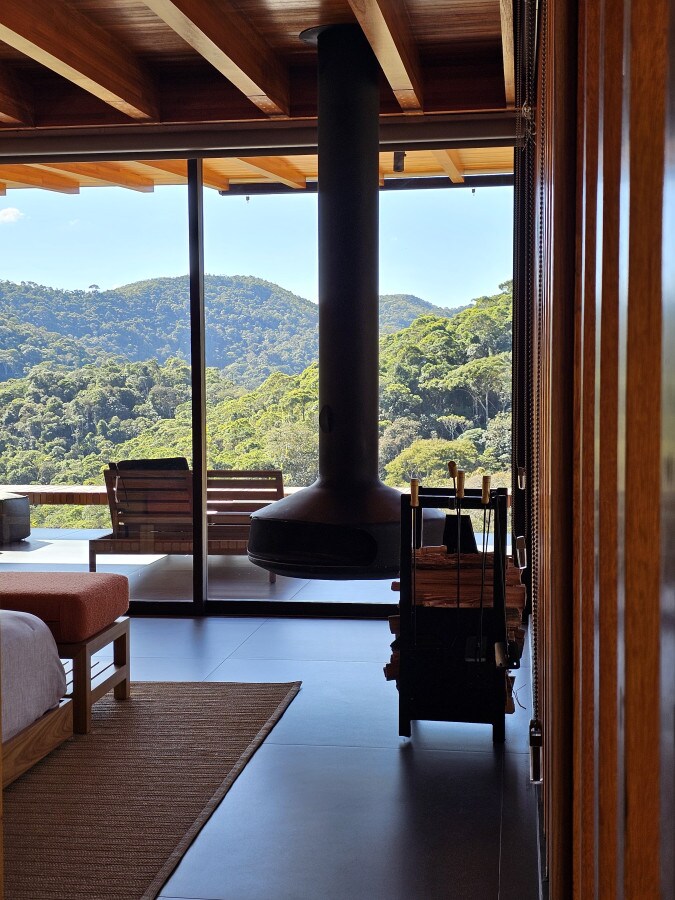 A modern interior is seen featuring a suspended black fireplace, positioned beside a wooden cart. Large glass doors open to reveal a stunning view of green mountains, while a wooden seating area is visible outside, complementing the serene ambiance.