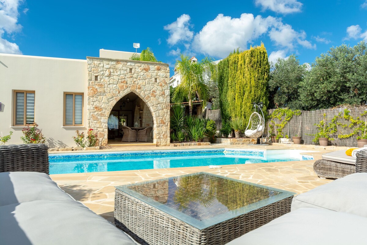 A serene outdoor space surrounds a private swimming pool, with stone walls and vibrant greenery in the background. Comfortable lounge chairs are positioned nearby, and a stylish glass-top table is seen in the foreground, reflecting the clear blue sky and lush surroundings.