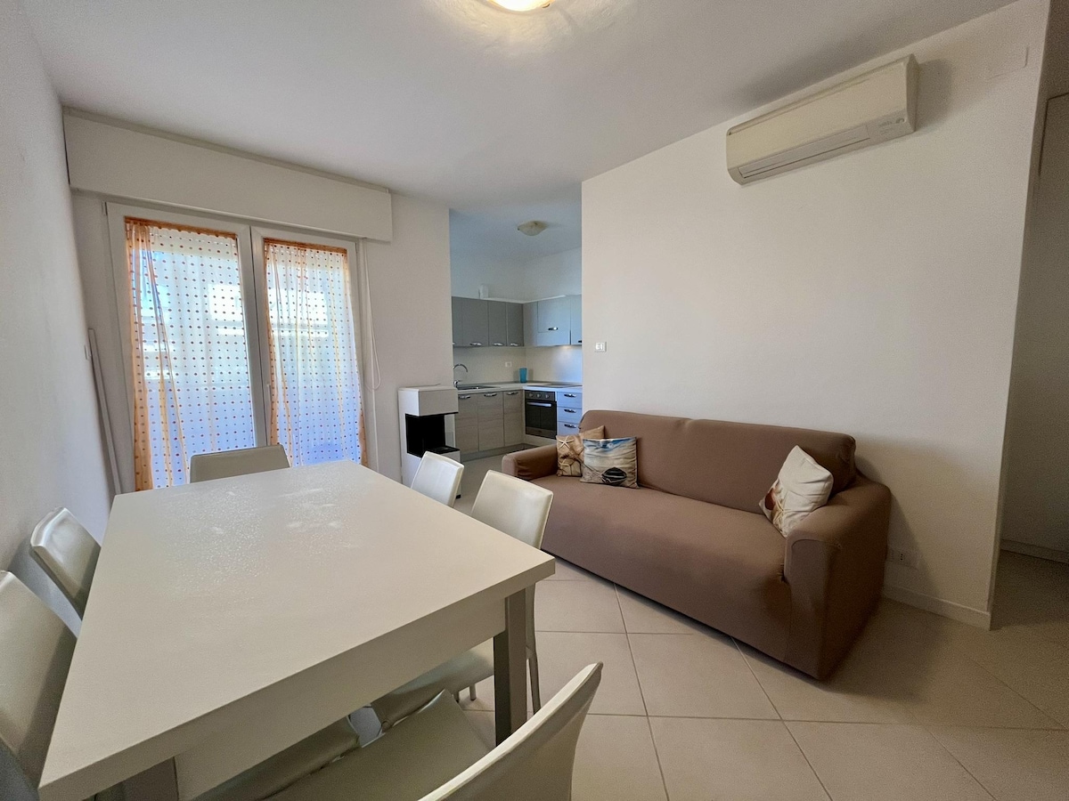 A bright living area features a beige sofa accompanied by a decorative pillow. A white dining table is surrounded by four chairs. Light filters in through dual glass doors leading to a terrace, and an air conditioning unit is mounted on the wall.