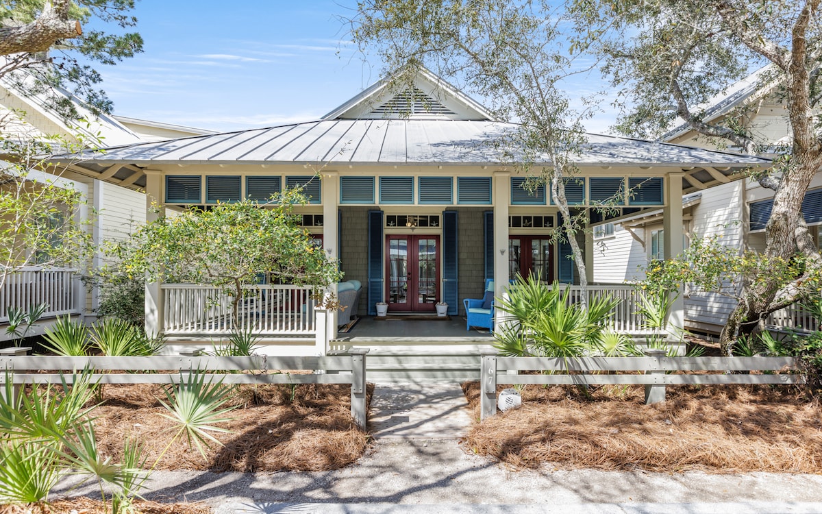 A charming single-story home is showcased with a metal roof and a wide front porch. Lush greenery, including palm plants, surrounds the entrance. The facade features large windows and several seating areas, inviting relaxation in the outdoor space.