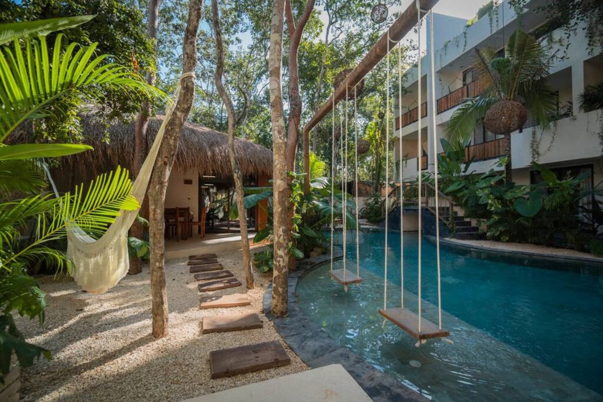 A serene outdoor space is framed by lush greenery, featuring a tranquil pool and natural stone pathways. Two wooden swings hang from a sturdy beam, inviting moments of relaxation, while a thatched-roof structure is visible in the background along with a dining area.