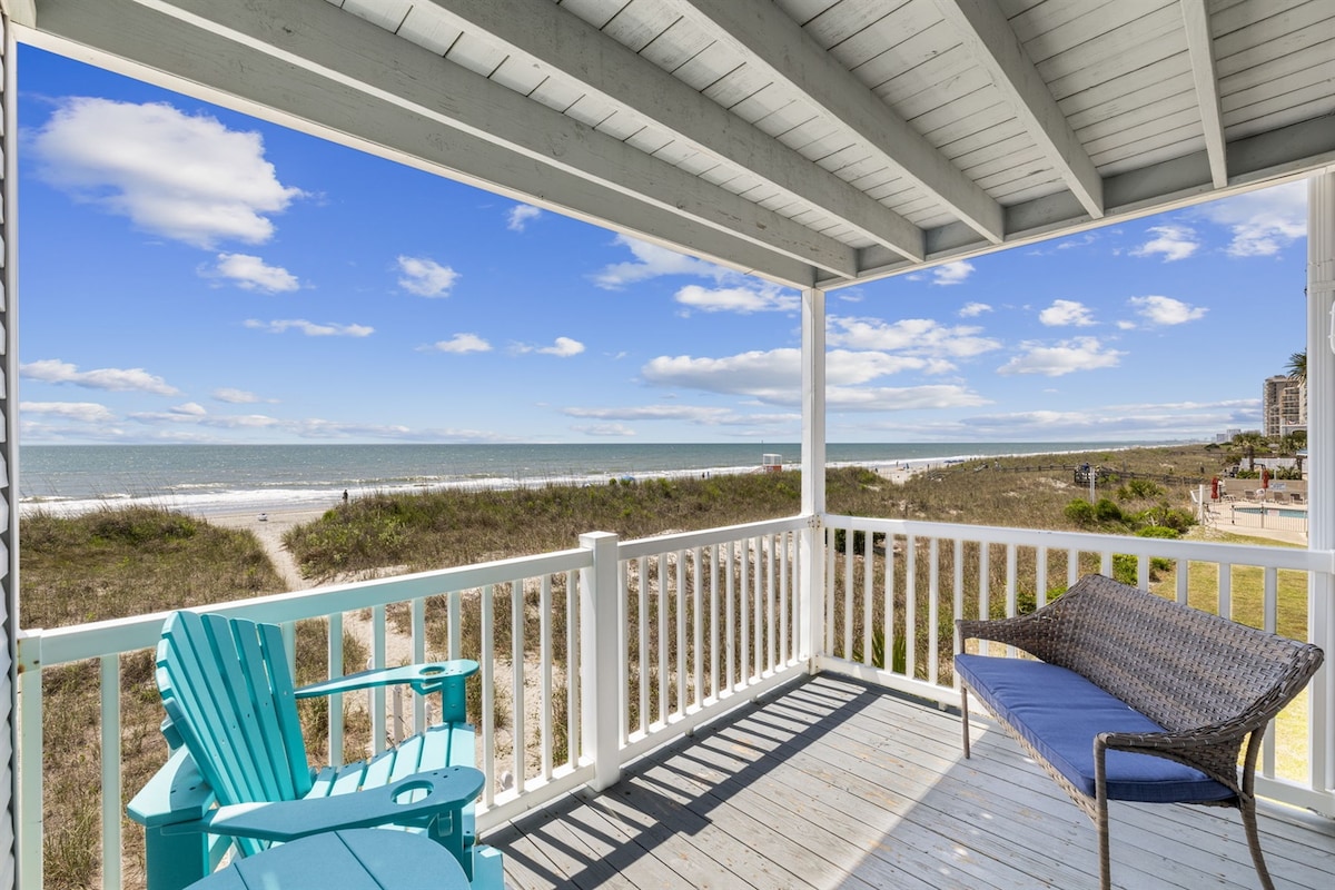 A spacious balcony with white railings overlooks the sandy beach and ocean. Two chairs are positioned for relaxation, allowing for ocean vistas and gentle sea breezes. The clear blue sky complements the serene coastal setting.