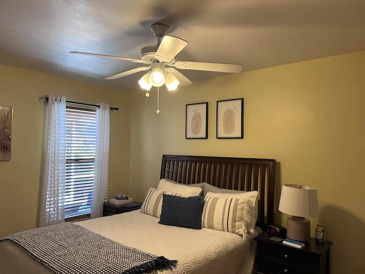 A bedroom featuring a double bed with patterned pillows and a throw blanket, flanked by wooden nightstands with a lamp and clock. A ceiling fan is above, and two framed artworks hang on the yellow wall.