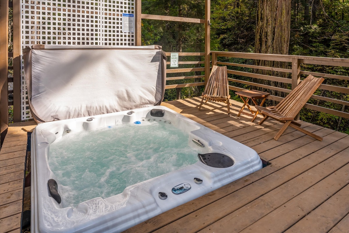 A hot tub is positioned on a wooden deck, surrounded by greenery and forest views. Two wooden lounge chairs are placed nearby, inviting relaxation in a tranquil outdoor setting.