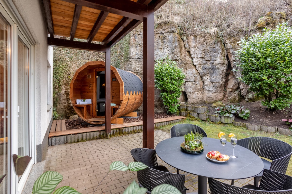A serene outdoor area features a round wooden sauna positioned against a natural rock wall, surrounded by greenery. A circular dining table with black chairs is set for guests, complete with a platter of fruit and drinks, inviting relaxation in a private garden setting.