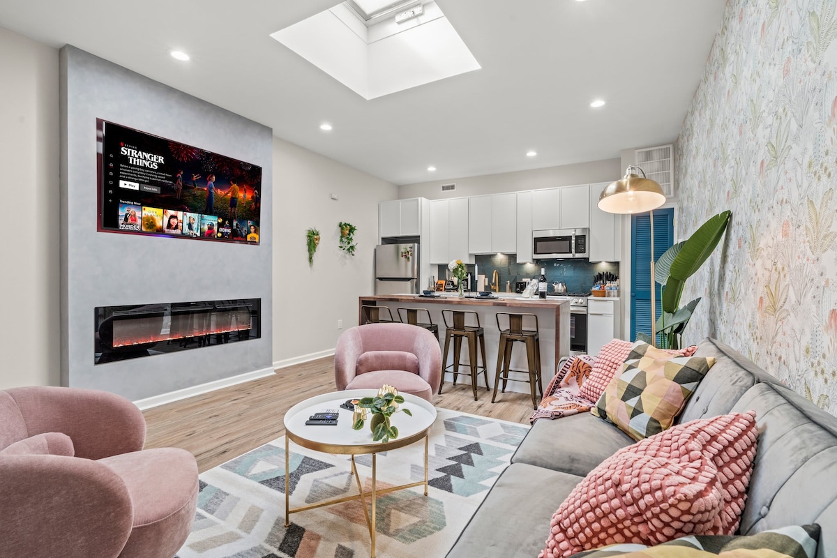 A modern living area features a cozy seating arrangement with plush sofas and armchairs. A sleek electric fireplace adds warmth, while a smart TV mounted above provides entertainment options. Natural light floods the space through a skylight, enhancing the airy feel of the open-concept layout.