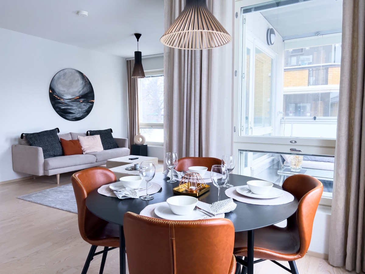 A dining area features a round black table surrounded by four tan leather chairs. A modern light fixture hangs above, while a grey rug complements the wooden floor. The room includes a plush sofa and a large window, allowing natural light to illuminate the space.