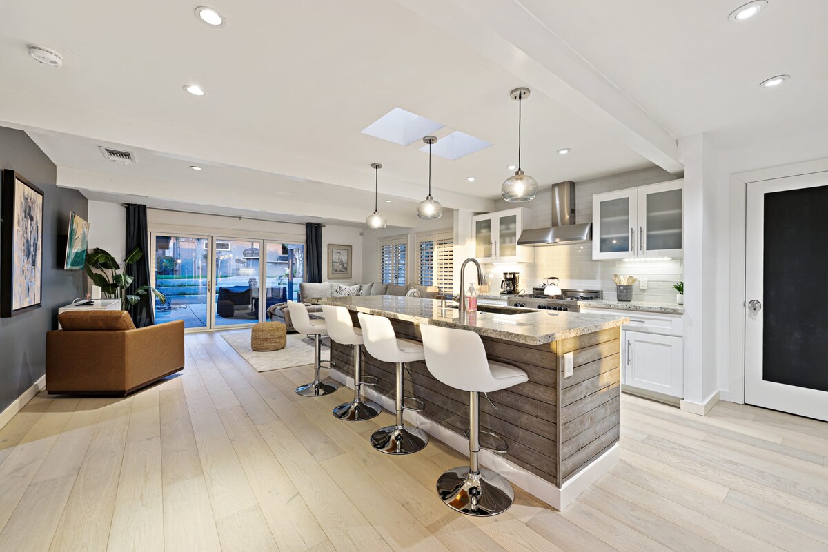An open-concept kitchen features a spacious island with four sleek bar stools. Modern cabinetry and stainless steel appliances add to the contemporary design, while large sliding glass doors lead to an outdoor area, allowing natural light to fill the space.
