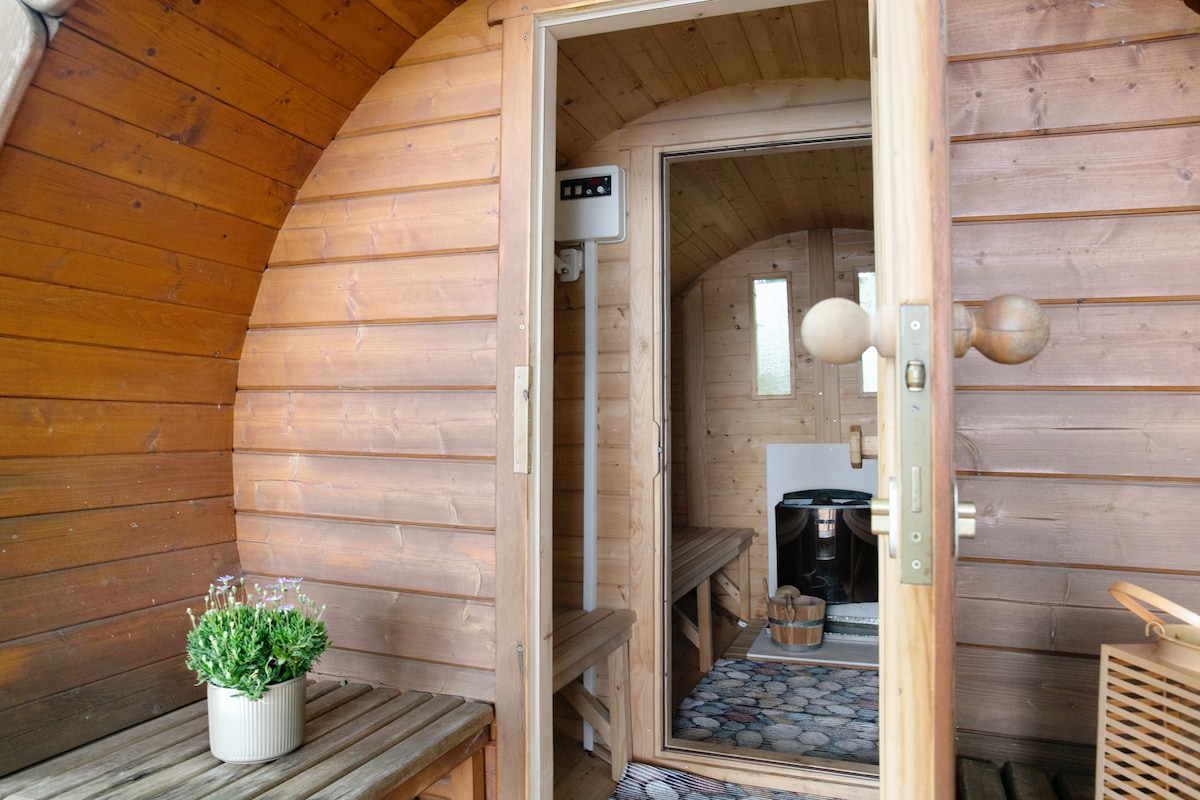 A wooden sauna is featured with a rounded structure that showcases natural wooden walls and benches. The entrance door is partially open, revealing a cozy interior. A small plant sits on a wooden bench, adding a touch of greenery to the inviting space.
