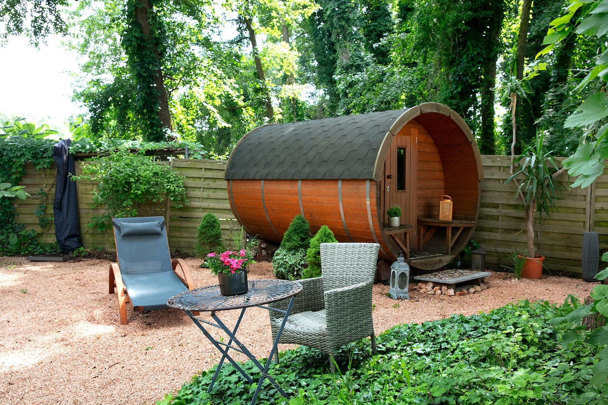 A wooden barrel sauna is nestled among lush greenery in a private outdoor area. A small round table with two chairs is positioned nearby, alongside a lounge chair. Decorative plants and gravel paths enhance the serene environment, while sunlight filters through the surrounding trees.
