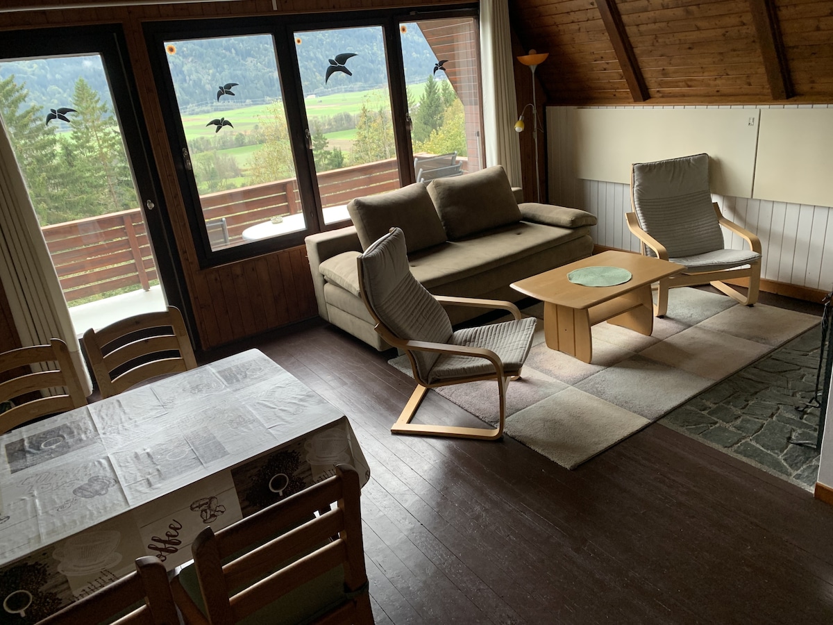 A comfortable living area features a plush sofa paired with two armchairs, arranged around a simple coffee table. Large windows provide a view of the surrounding landscape and allow natural light to fill the space. A dining table is positioned nearby.