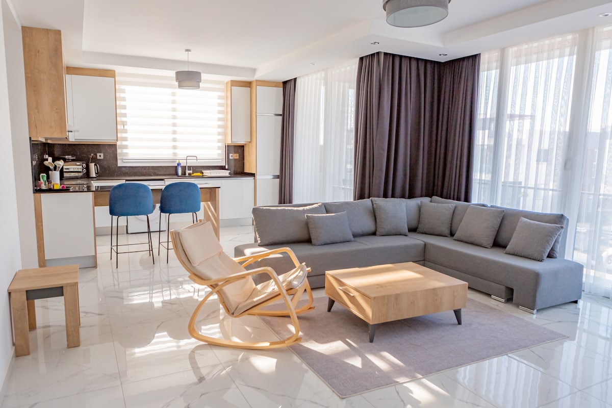 A modern living area features a spacious gray sectional sofa and a wooden coffee table on a neutral rug. Natural light filters through sheer curtains, illuminating the open kitchen with bar stools. A light-colored rocking chair is positioned nearby, enhancing the inviting and relaxed ambiance.
