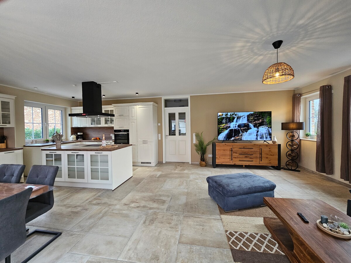 An open-concept living area is displayed, featuring a modern kitchen with sleek cabinetry and a large island. A cozy seating area includes a dark-blue ottoman and a brown table, with a large flat-screen TV mounted on a wall. Natural light filters in through the windows.