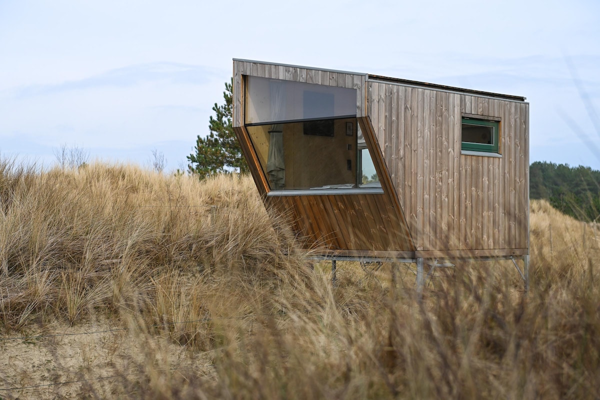 A modern tiny house is set among golden grasses, showcasing a distinctive angled design with large windows. The wood exterior reflects natural tones, while the structure is elevated above the ground. The surrounding landscape features gentle dunes and sparse trees in the background.