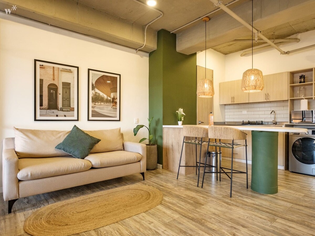 A modern living area features a light-colored sofa with a decorative pillow, complemented by a natural fiber rug. Two framed prints adorn the wall, while a spacious kitchen area includes wooden cabinetry and pendant lighting. A small dining table with stools is positioned nearby.