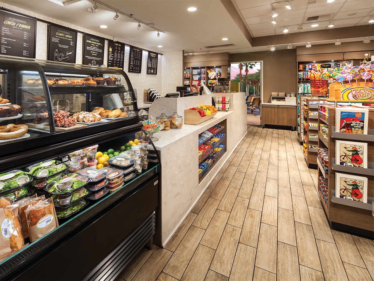 A welcoming convenience store features a glass display case filled with assorted fresh pastries and snacks. Shelves stocked with various packaged goods line the walls, while brightly colored candy and snack items provide visual appeal. Warm lighting enhances the inviting atmosphere of the space.