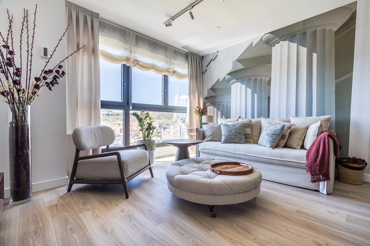 A bright living area features a light-colored sofa adorned with decorative cushions, alongside a modern armchair. A round ottoman sits in front, and a small table is positioned centrally. Large windows offer ample natural light, with soft drapes complementing the design.