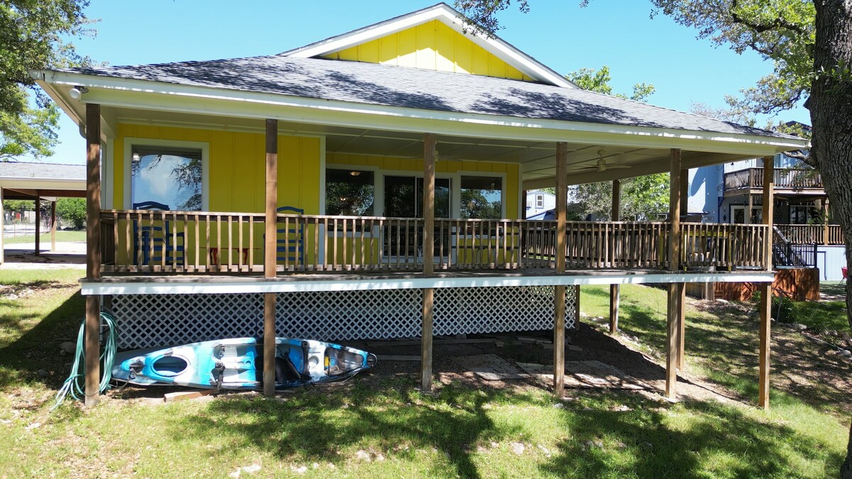 A bright yellow house is highlighted by a spacious covered porch supported by wooden posts. Below the porch, two kayaks are placed on the grass, indicating opportunities for lakeside activities. The surrounding area is filled with trees, providing shade and a natural setting.