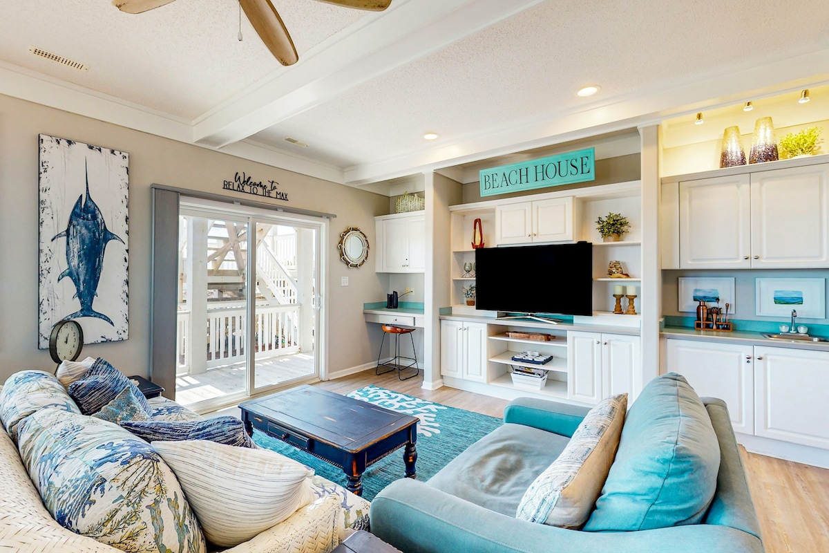 A bright living room is enhanced by natural light from sliding glass doors leading to a deck. Plush seating in coastal colors and patterns surrounds a wooden coffee table. A large TV is mounted on a white entertainment center with decorative items, contributing to a relaxed beach house ambiance.