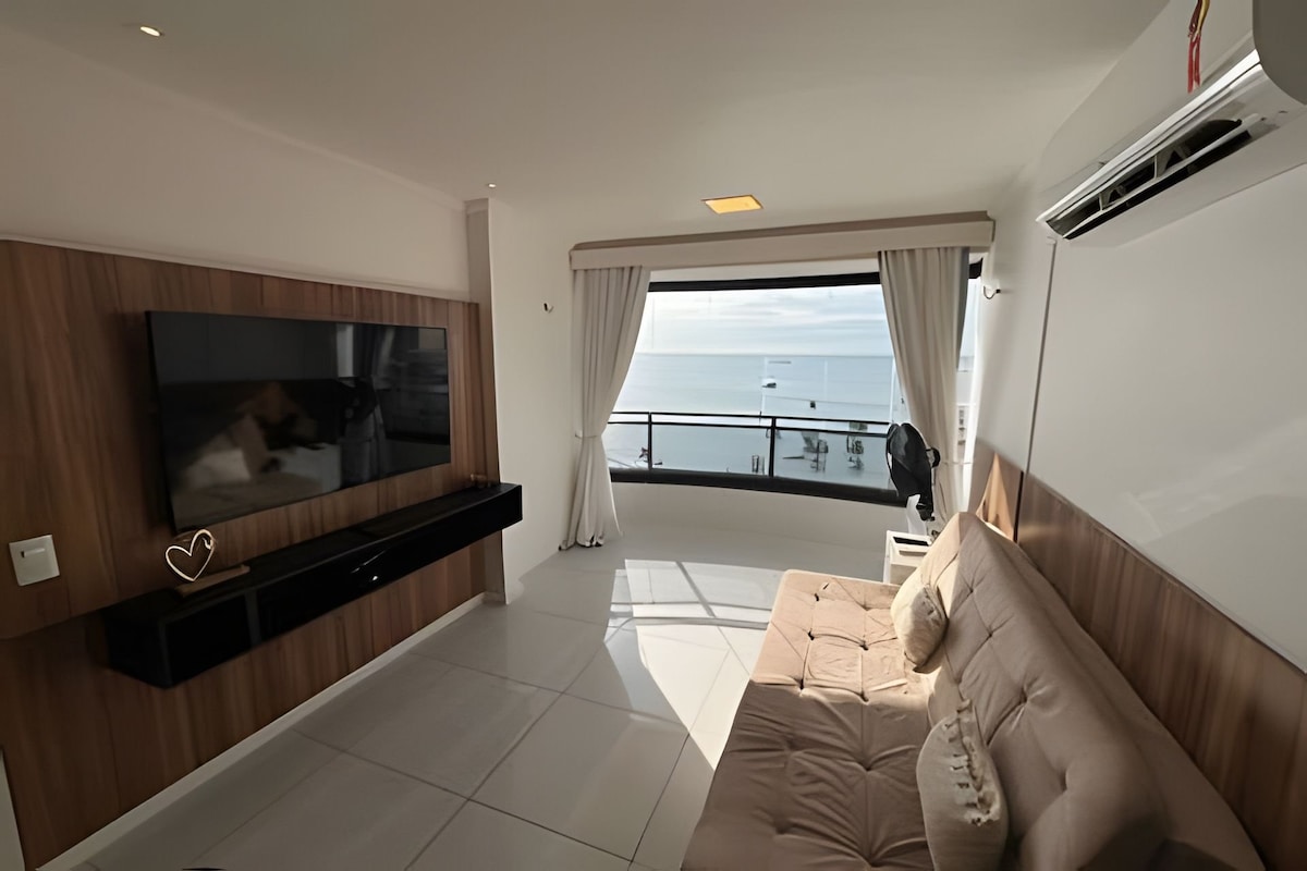 A comfortable living area features a large flat-screen TV mounted on a wooden wall. Light-colored curtains frame the panoramic view of the sea through the glass sliding doors. A cozy sofa bed is positioned for relaxation, with sunlight creating a bright ambiance.
