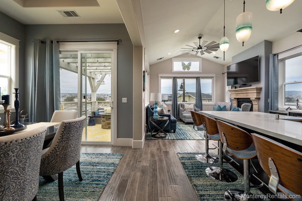 An open-concept living space features a modern kitchen and dining area with bar seating. Large windows frame scenic views, while a comfortable seating arrangement includes a large sectional with decorative pillows. A bright rug enhances the ambiance, and a fireplace adds a touch of warmth.