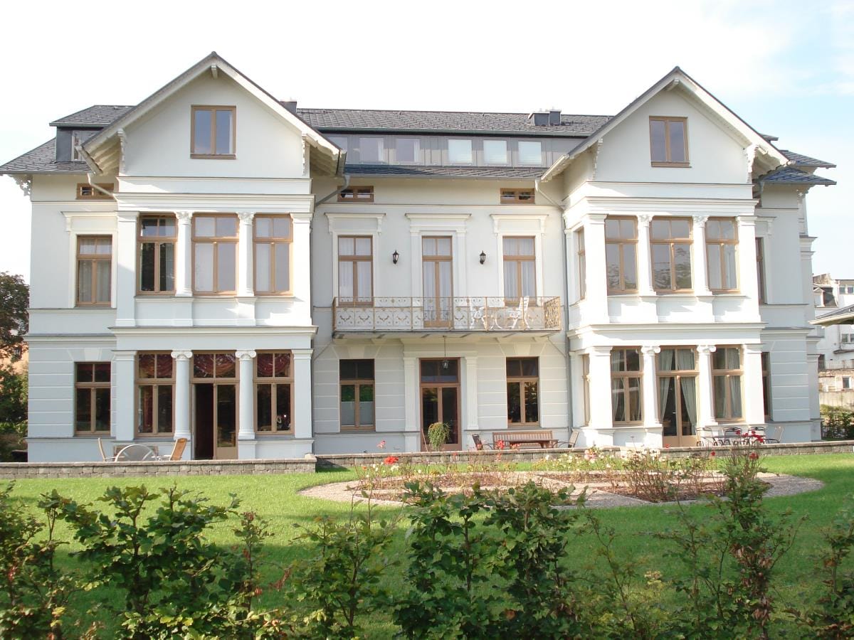 The building features a grand façade with multiple large windows and a balcony. The structure showcases classic architectural details, surrounded by a well-maintained garden with low hedges and colorful flower beds. The exterior reflects a welcoming atmosphere with its light-colored walls.