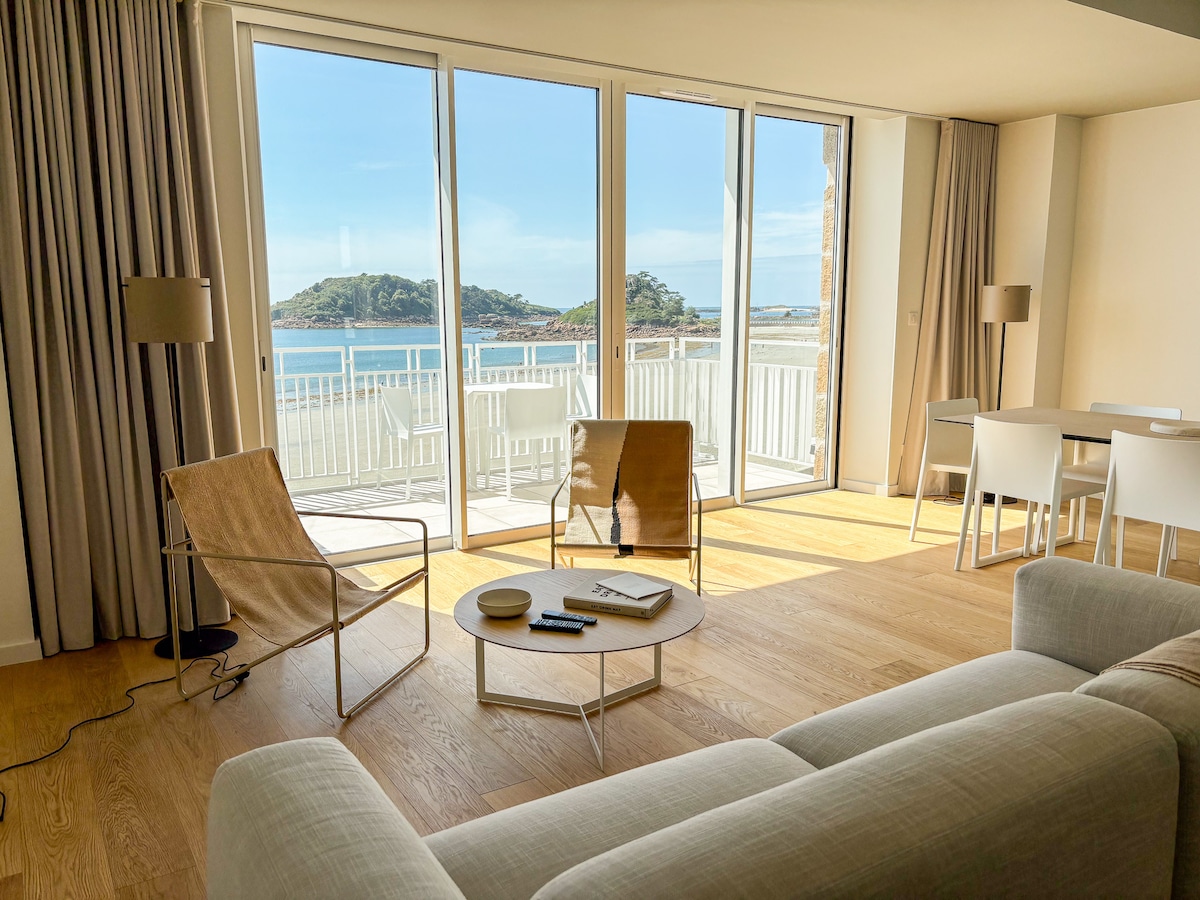 A spacious living area is showcased with large glass doors providing panoramic views of the sea. Two modern chairs and a circular coffee table are positioned in front of a comfortable sofa. Light wood flooring complements the serene atmosphere, creating a welcoming space.