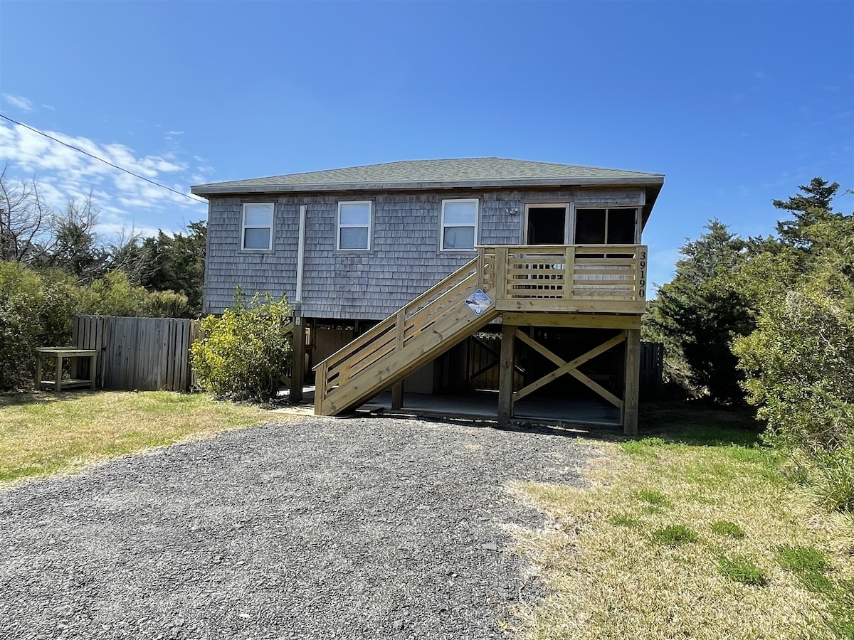 A raised cottage is showcased, featuring a wooden ramp leading up to a front deck. Shingle siding and a green roof complement the natural surroundings, while gravel leads to the home. Lush greenery and a fenced area are visible nearby, enhancing outdoor space.