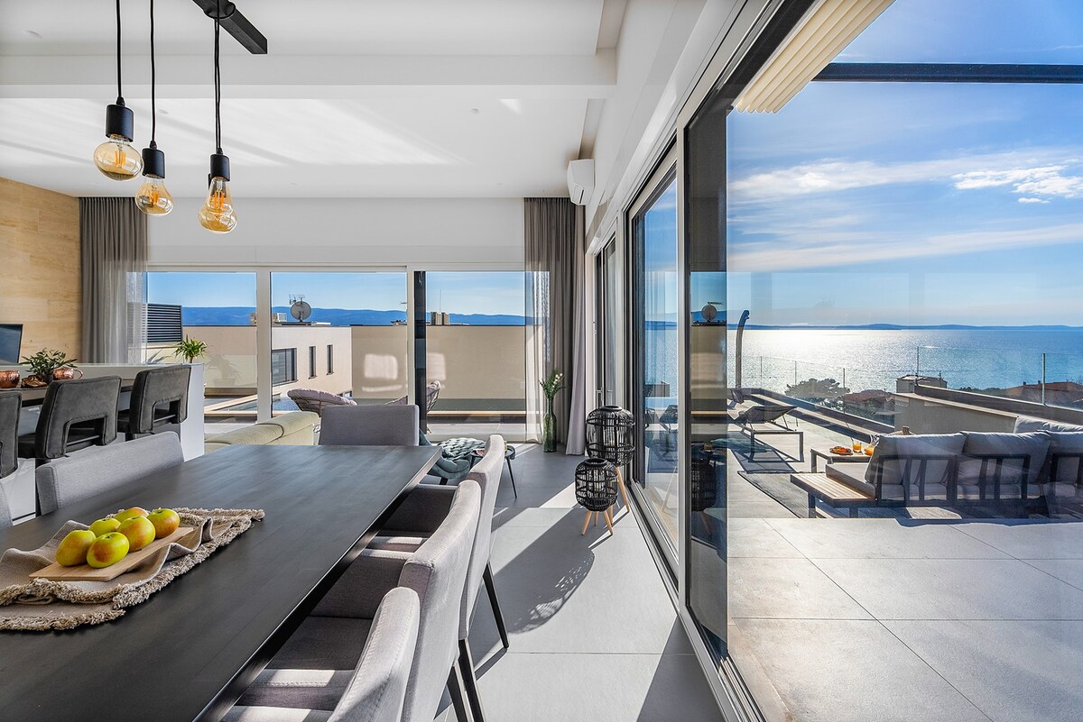 The open-plan dining and living area is designed with large glass walls, offering panoramic sea views. A dark dining table is set for eight. Warm sunlight floods the space, enhancing the connection between the interior and the outdoor terrace.