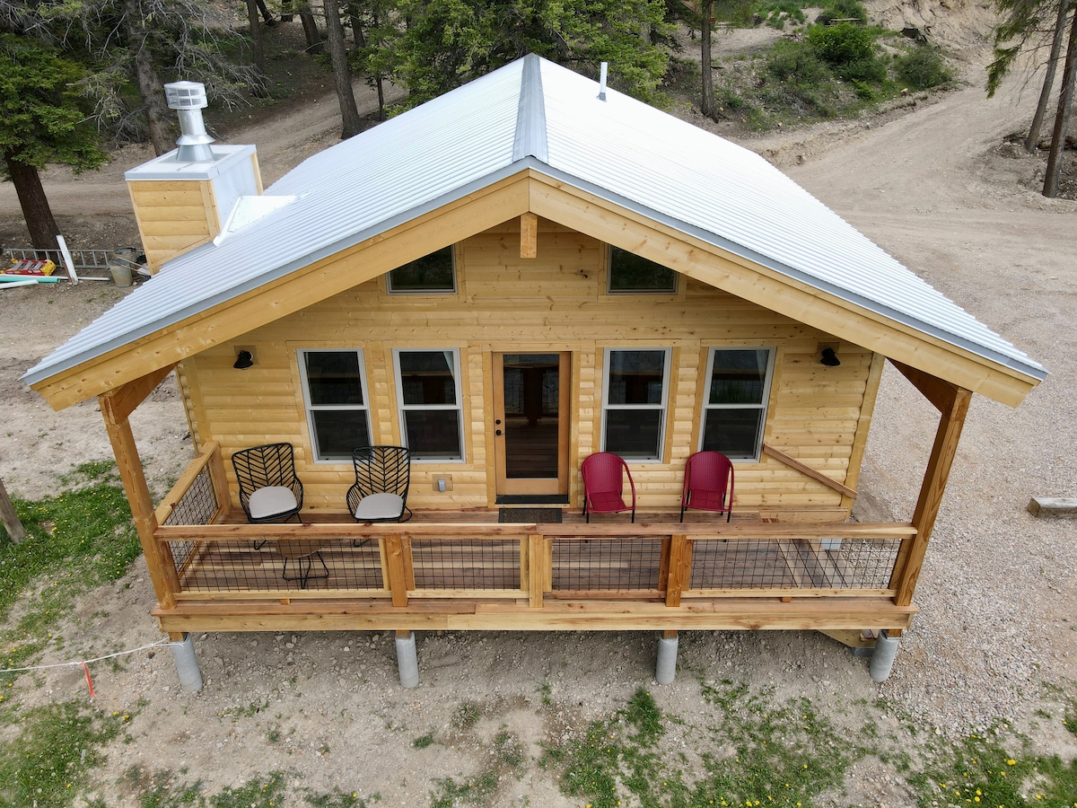 The cabin features a wooden exterior with a metal roof, showcasing a spacious front porch. Two outdoor chairs are positioned on the porch, offering a space to relax. Large windows allow natural light to flood the interior, enhancing the welcoming atmosphere of the home.