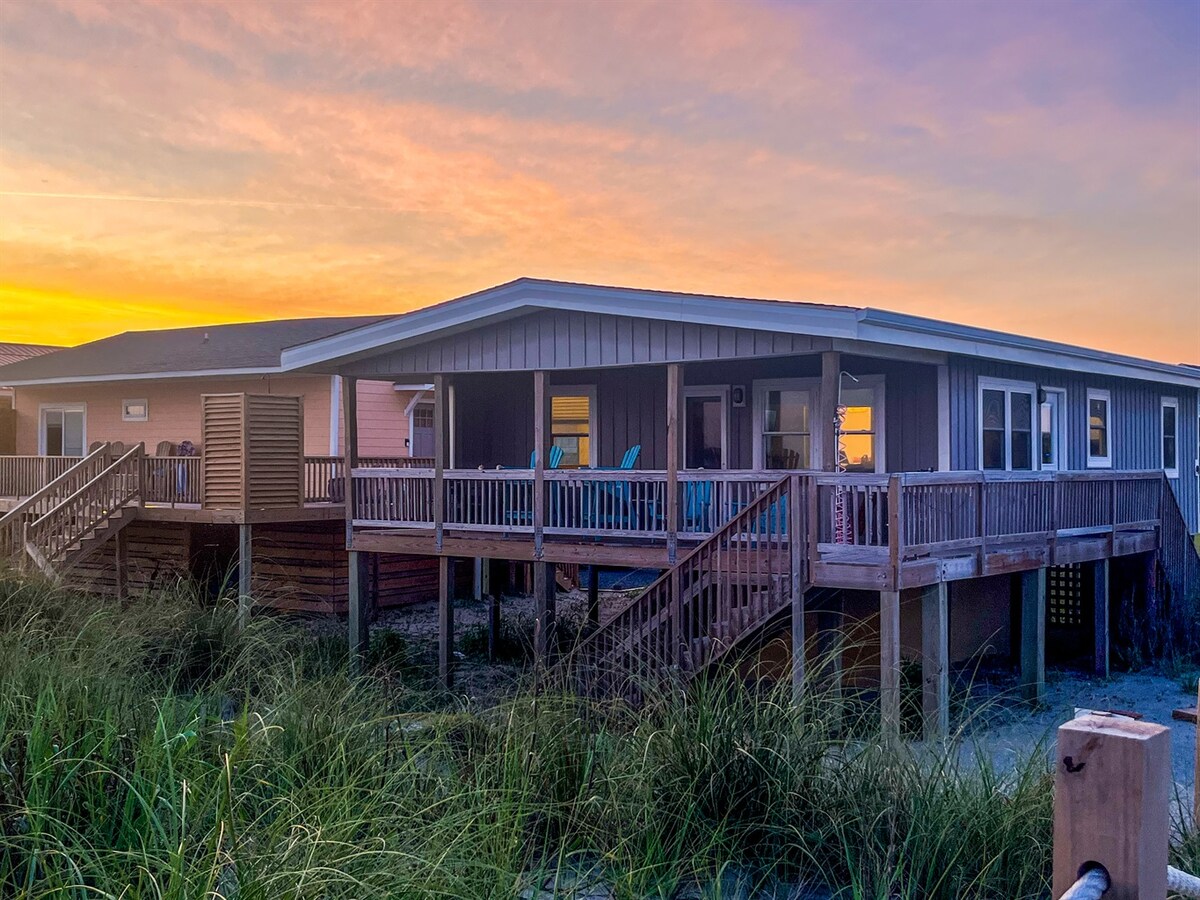 The beach front bungalow is showcased against a colorful sunset, highlighting its wooden exterior and expansive deck. Outdoor seating is positioned to take in the views, and surrounding tall grass offers a natural setting that complements the coastal environment.