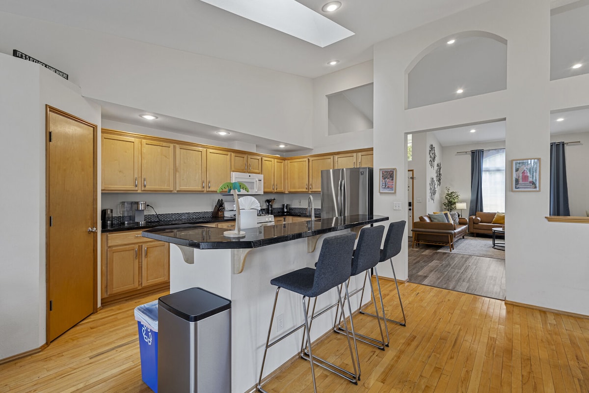 The spacious kitchen features wooden cabinetry, a large island with seating for three, and stainless steel appliances. Natural light streams in through the skylight, enhancing the open layout connected to the living area. A trash can and recycling bin are placed conveniently nearby.
