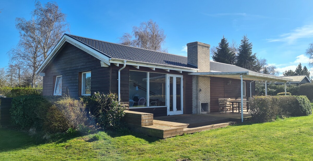 The exterior of the summer house features a wooden facade complemented by a spacious terrace. A fireplace is located on the side of the house, while large glass doors allow for natural light to brighten the interior. Surrounding greenery enhances the tranquil setting.
